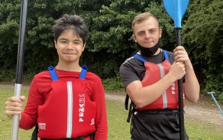 Max and Jack standing together with red life vests on and a paddle each which has a blue end. They're standing on a section of grass with tall green trees in the background, very scenic!