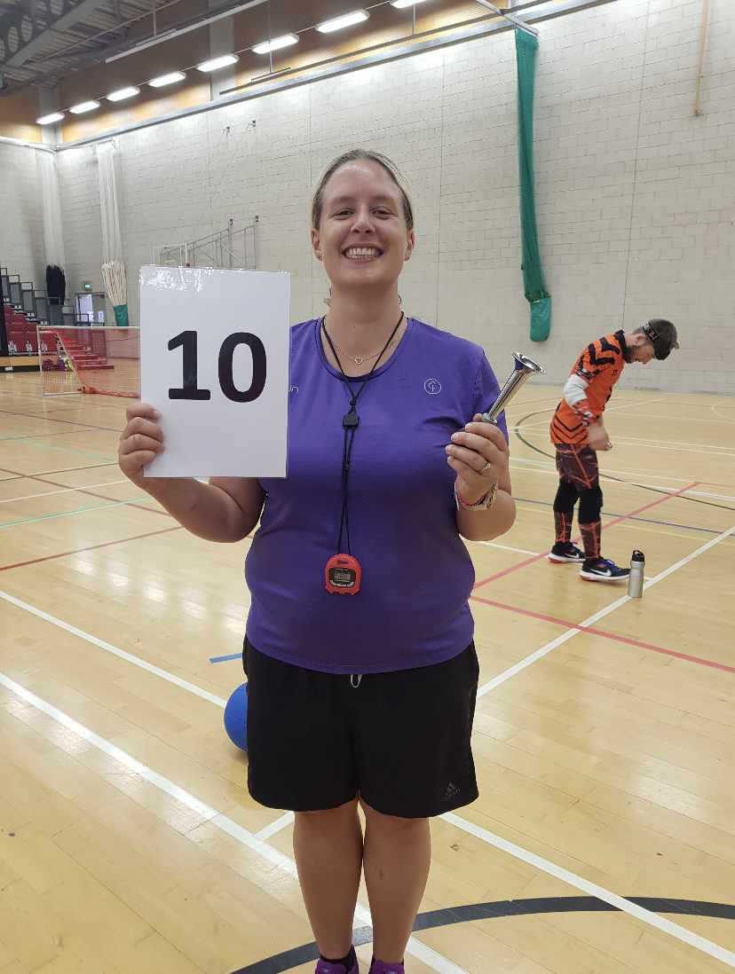 Kirsty Mills standing in a purple t-shirt and black shorts, holding up a laminated piece of paper with '10' on it, signifying 10 seconds. In the other hand Kirsty is holding a hooter.