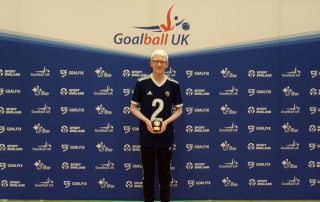 Leo Stubbs standing with his top Mae goalscorer trophy after winning it in the 2021/2022 Novice season. Leo is standing proudly with his plaque with a Goalball UK banner behind him.