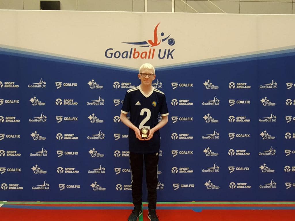 Leo Stubbs standing with his top Mae goalscorer trophy after winning it in the 2021/2022 Novice season. Leo is standing proudly with his plaque with a Goalball UK banner behind him.