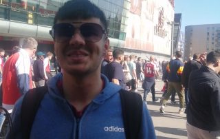 Mustafa Abbas outside the Emirates stadium ahead of an Arsenal game. Mustafa has a bright smile whilst wearing a blue hoodie and dark, cool looking sunglasses!
