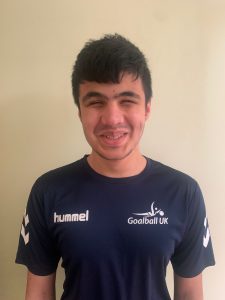 Mustafa Abbas standing in front of a cream wall in his navy blue #FindTheNext Goalball Academy shirt, with a big cheesy smile!