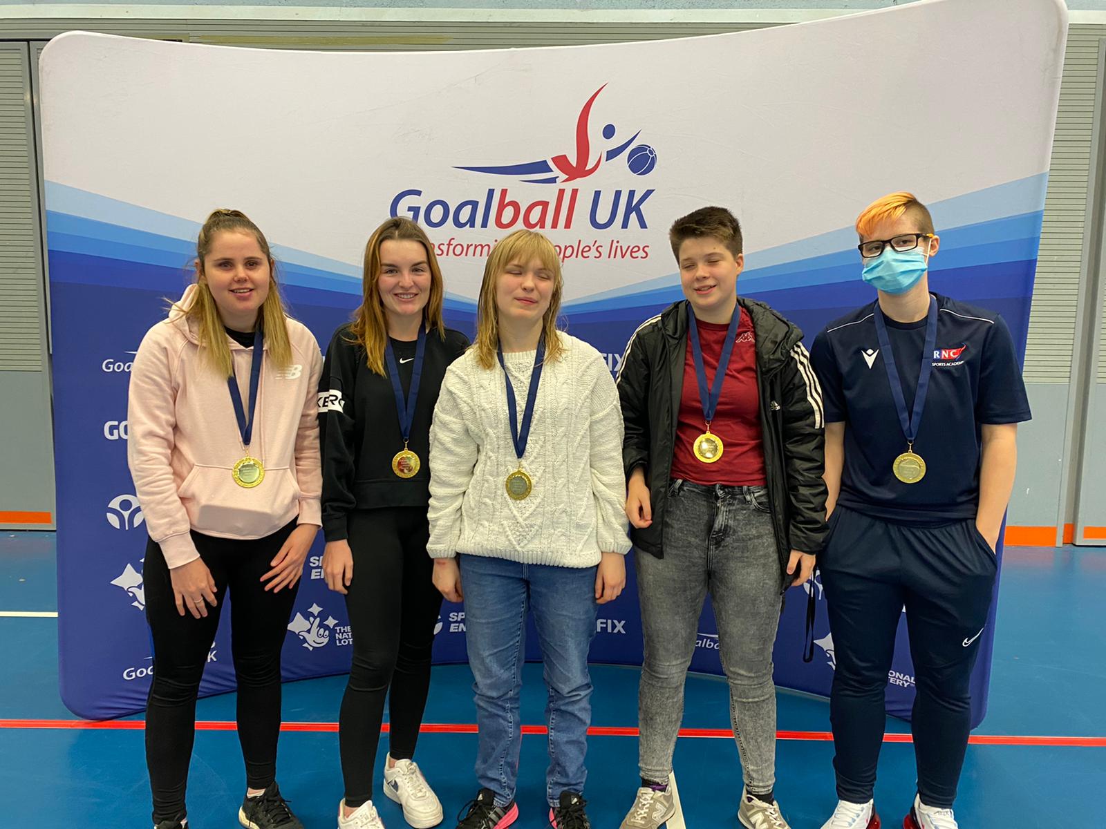 RNC Ladies regional team standing for their post competition gold medal photo. 4 players are in casual clothing with their coach Meme Robertson who is wearing a navy blue RNC training tracksuit.