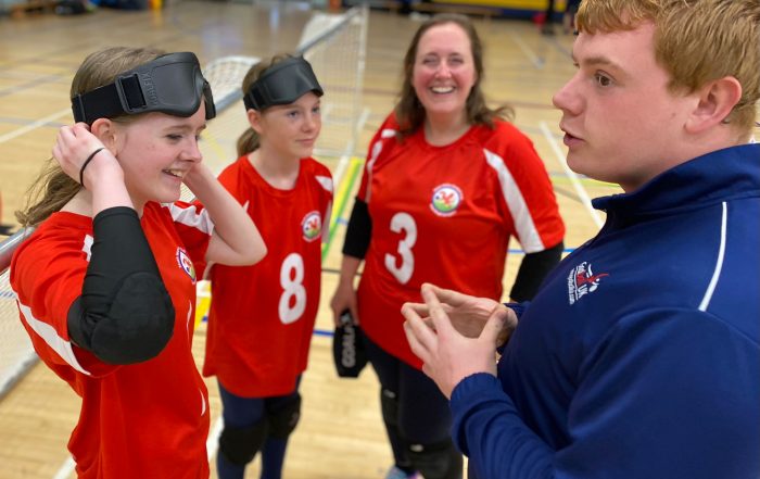 Three female South Wales goalball team members stand talking to Chris. He is wearing a blue Goalball UK top. A purple square with the 'This Girl Can' logo is in the bottom left of the image.