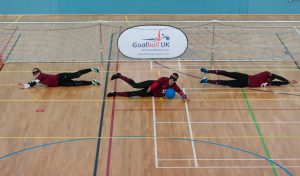 Three goalball players on the floor infront of the goal in defence.