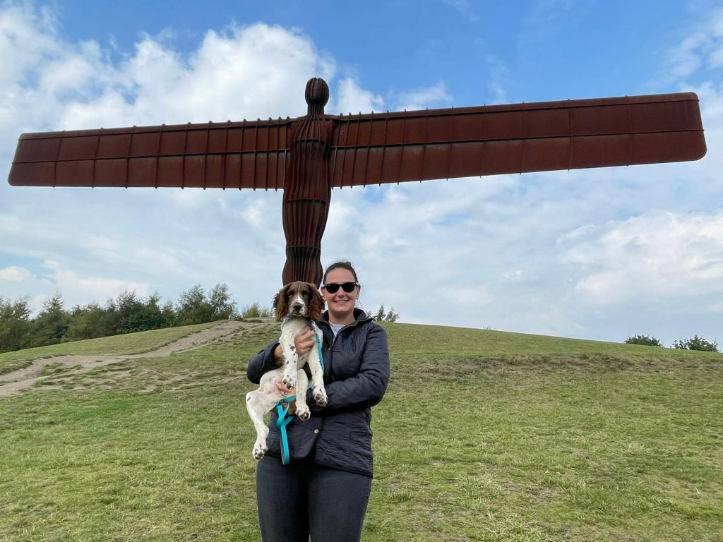 Dani Dillon stood under the Angel of the North statue holding a dog. Dani is wearing a black coat and sunglasses.