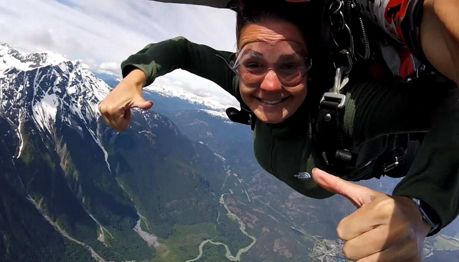 Dani Dillon skydiving, she is looking at the camera wearing goggles and smiling holding two thumbs up