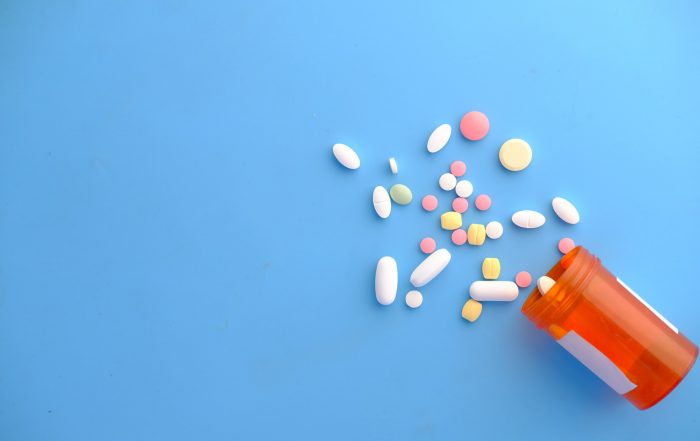 Photo of a pill bottle on it's side with different shaped and coloured tablets falling out of the bottle. The background is blue.