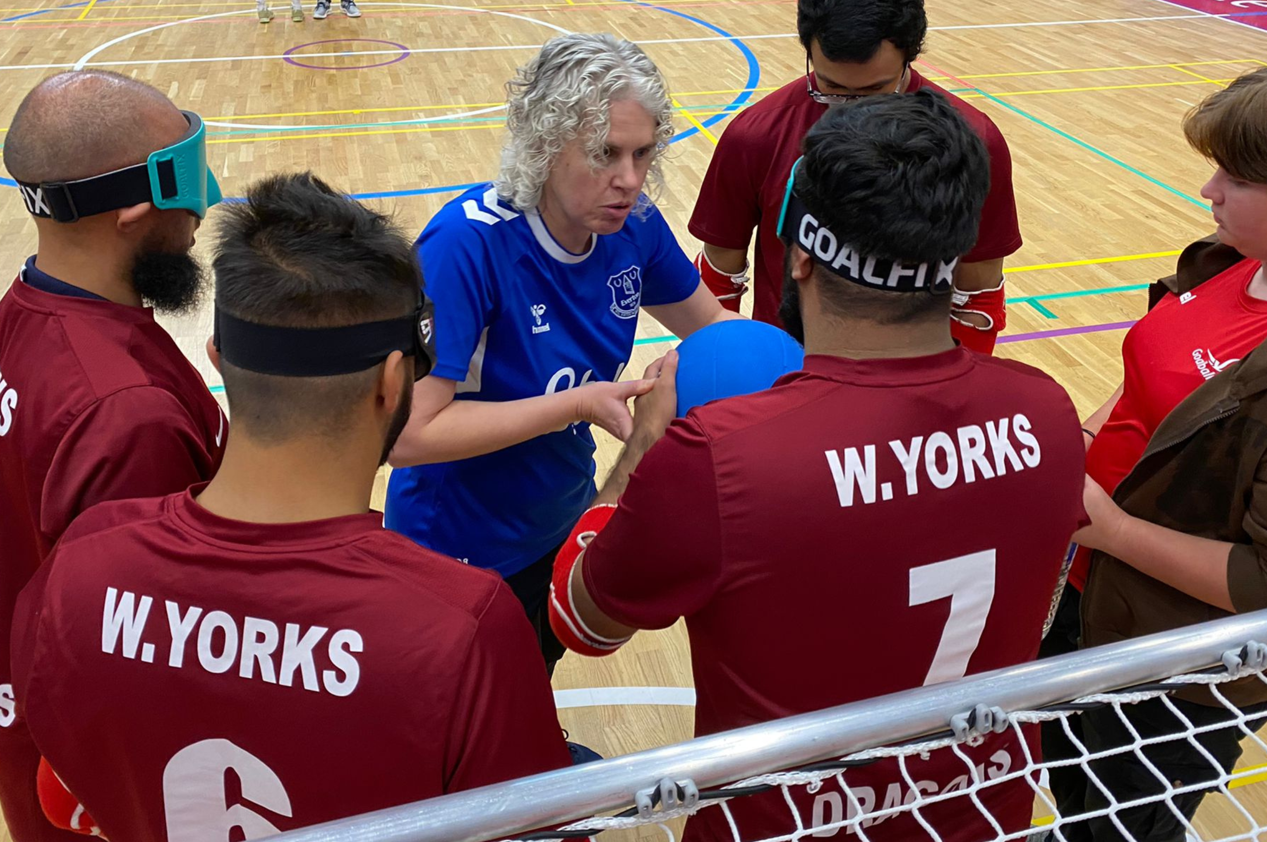 Kathryn Fielding speaks to Bradford Goalball Club team members during a team huddle. Kathryn is passing the Goalball to a player who is wearing eye shades