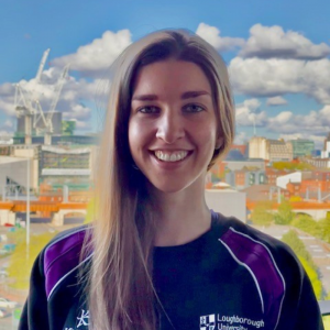 Anna Martin smiling in a dark blue and purple Loughborough jumper, with her hair, parted to her left. Behind Anna, are buildings within the Loughborough campus, and a partly cloudy, but bright blue sky.
