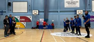 Photo of two goalball teams lined up before the start of the game. The photo is taken from a distance but they are all smiling at the camera