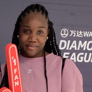 Emerlyne standing in front of a dark grey backdrop with the athletics Diamond League logo. Emerlyne is wearing a pink Adidas jumper and has an orange foam finger to show she’s a number 1 fan!