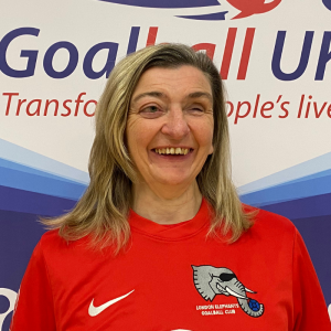 Tracy Compton stands in front of a blue and white Goalball UK banner which shows logos such as Goalfix, National Lottery, and Goalball UK. Tracy is smiling in her bright red London Elephants jersey.