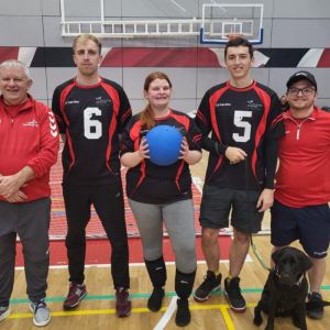Aaron and four other Goalball players from York St John Goalball Club are posing for a team photo. A woman in the centre is holding up a blue goalball. They are all wearing black and red kit.