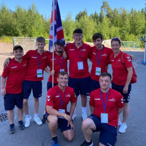 Group photo from the European Youth Paralympic Games with Aaron and others in red Goalball UK kit. The team are holding up a Union Jack flag and smiling for the camera.