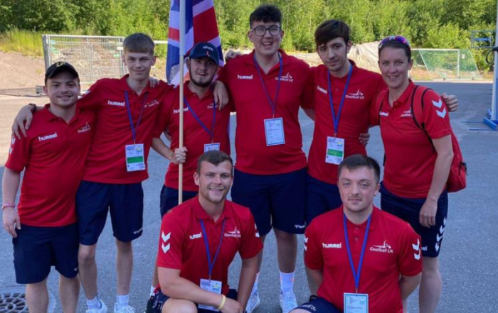 Group photo from the European Youth Paralympic Games with Aaron and others in red Goalball UK kit. The team are holding up a Union Jack flag and smiling for the camera.