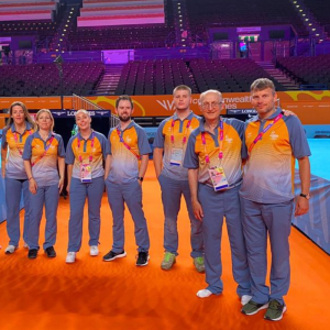 A group photo of Aaron Mitchel and other officials stood in a sports venue. They are all wearing great and orange kit and smiling for the camera.