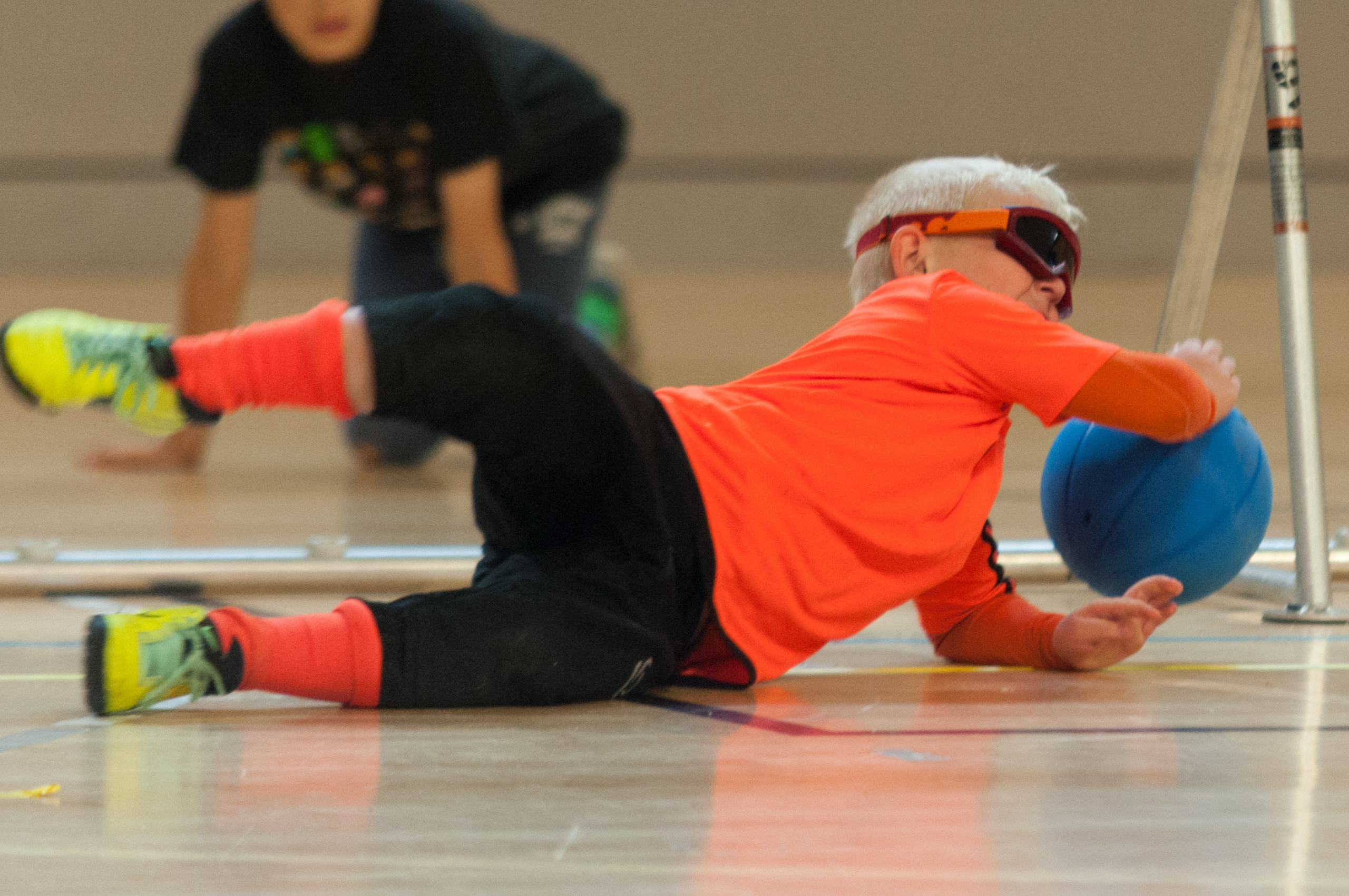 A child is on the floor of the court, stretched out to try and save a goal. He is wearing eye shades and a neon orange top and socks.