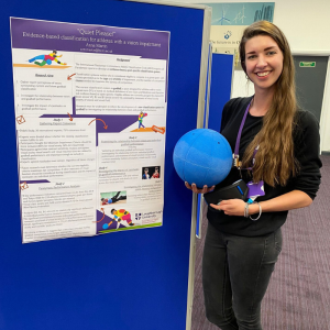 Anna stood holding a goalball in front of her academic poster