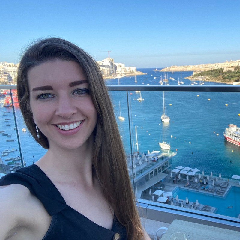 Anna smiling for a selfie stood on a balcony with a sunny harbour view in the background