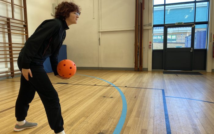 A slim dark haired woman gets ready to throw a junior goalball which is orange