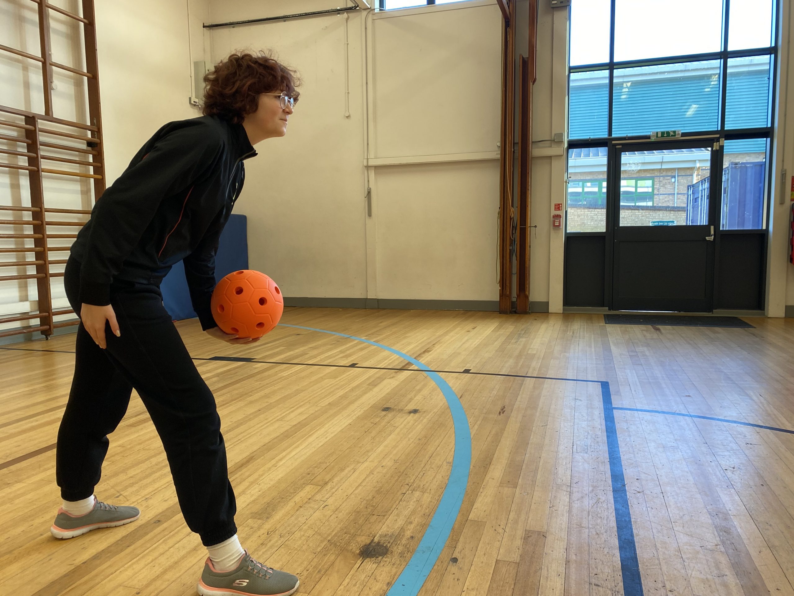 A slim dark haired woman gets ready to throw a junior goalball which is orange