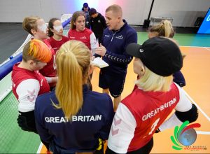 Aaron giving the GB Women's squad feedback post game at the 2022 Goalball World Championships. Players, coaches, and staff are standing on court in a circle in their GB kit.