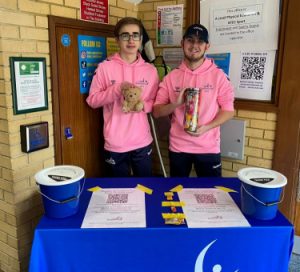 Harry Bainbridge and Jacob Hare of the Goalball UK Youth Forum standing together at their fundraising table. Harry is on the left holding a teddy bear, while Jacob on the right is holding a jar of sweets, their two fund-raising games!