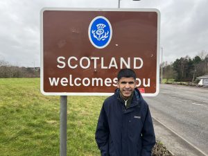 Yusuf Huraira standing in front of a brown sign which reads in white text "Scotland welcomes you". Yusuf is wearing a warm navy blue coat, in front of overcast skies.