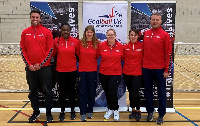 6 newly qualified goalball referees standing together in their red Goalball UK referee jackets. Left to right, Robert, Emerlyne, Magda, Leanne, Maggie, and Christopher, who are all stood in front of a Goalball UK banner.
