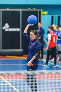 Coach Faye Dale holds a goalball above her head to throw it whilst holding a whistle and shouting