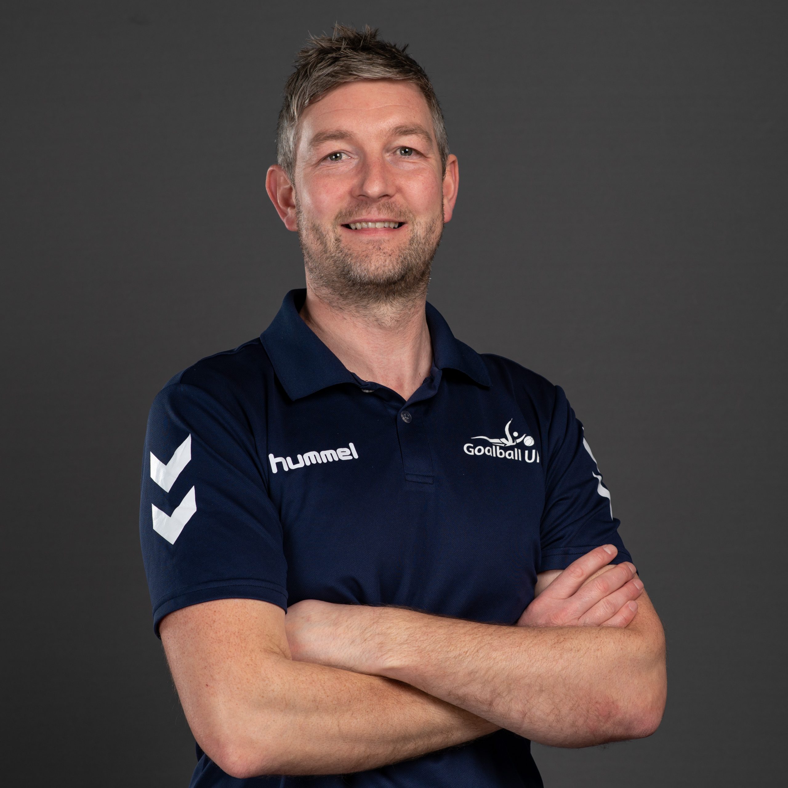 Gary Fraser headshot. Gary is wearing a dark blue Goalball UK t-shirt and is stood with arms folded against a dark grey background.
