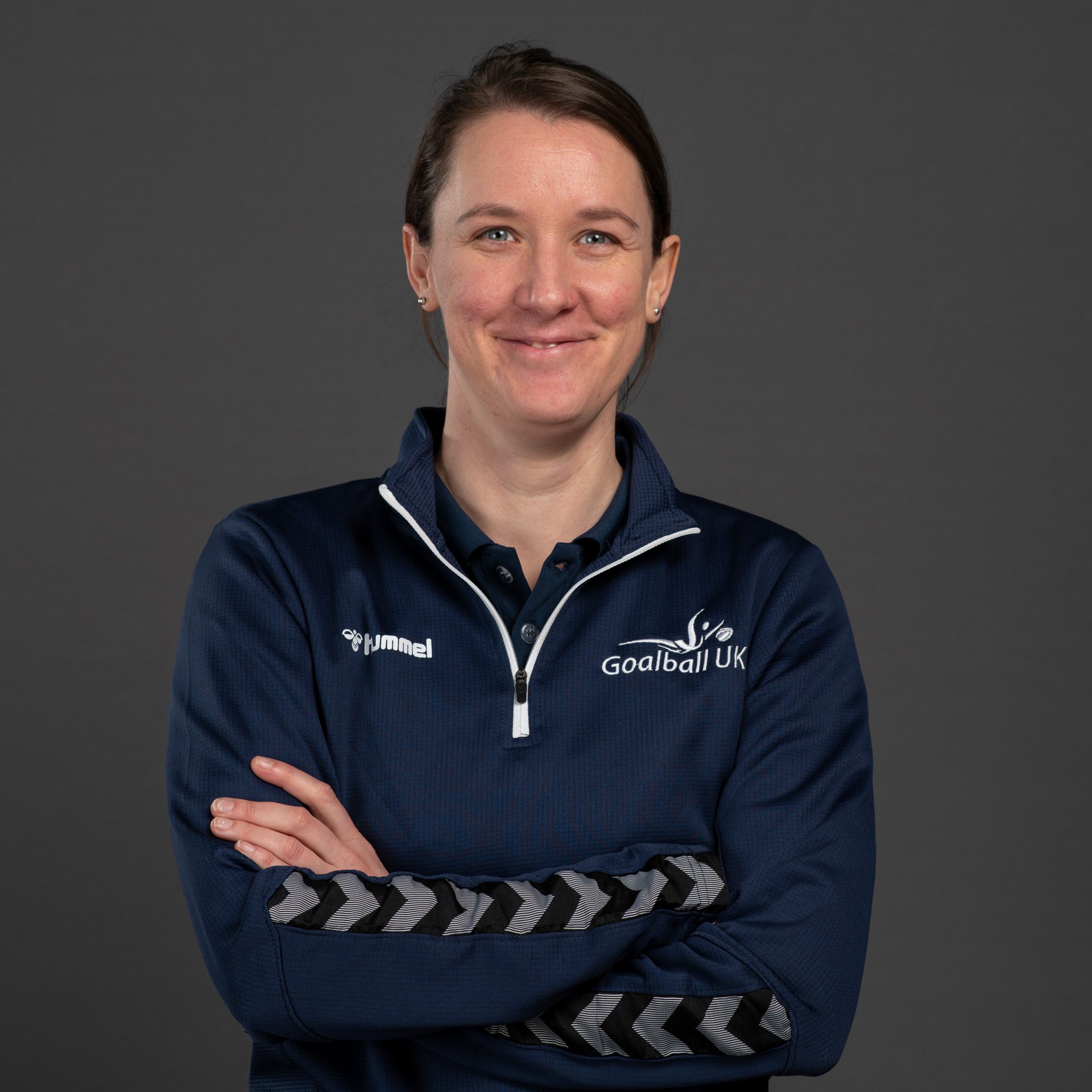 Faye Dale headshot. Faye is wearing a dark blue Goalball UK jumper and is stood with arms folded against a dark grey background.