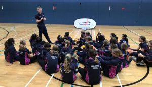 A school group sit on the floor of a sports hall and listen to Stephen Newey