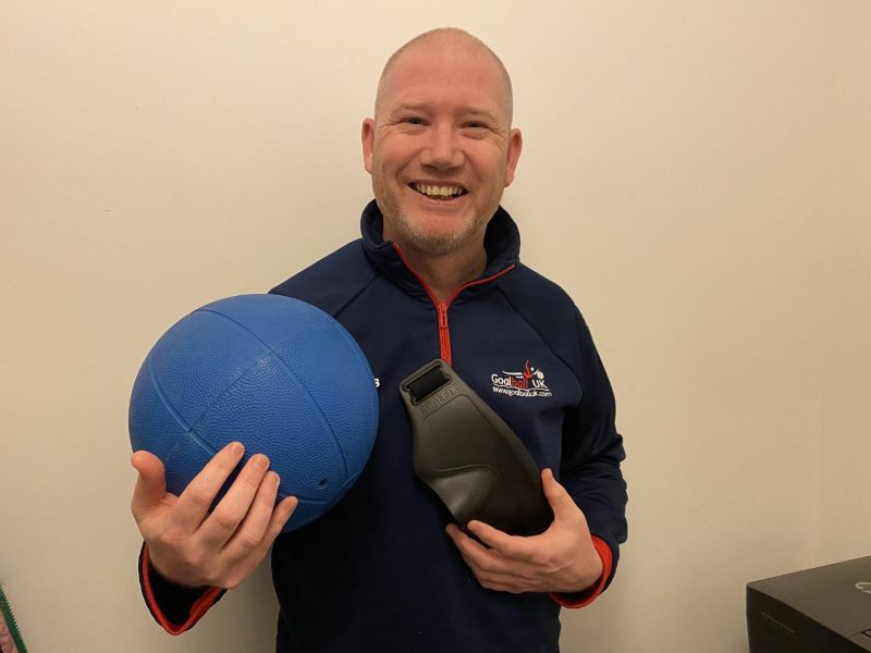 Mark Winder is stood holding a blue goalball and black eyeshades. He is smiling for the camera and wearing a blue Goalball UK top.