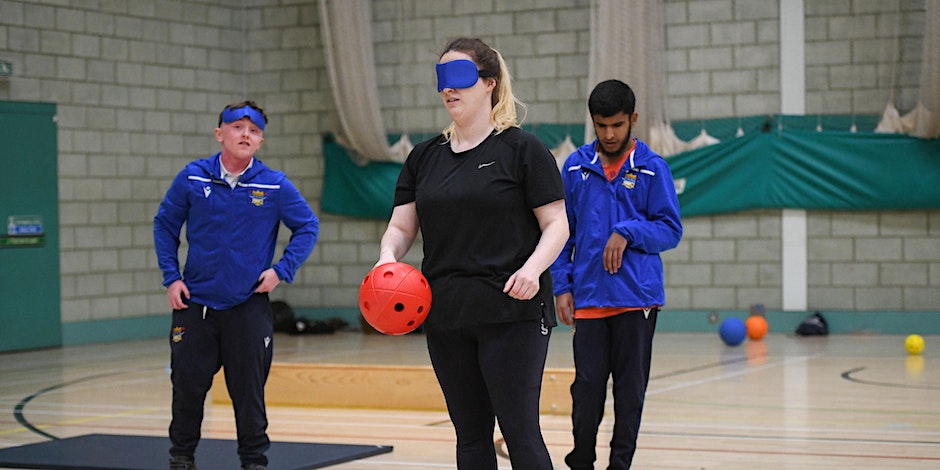 A woman is stood wearing soft blue eyeshades and holding a soft orange goalball during a taster session