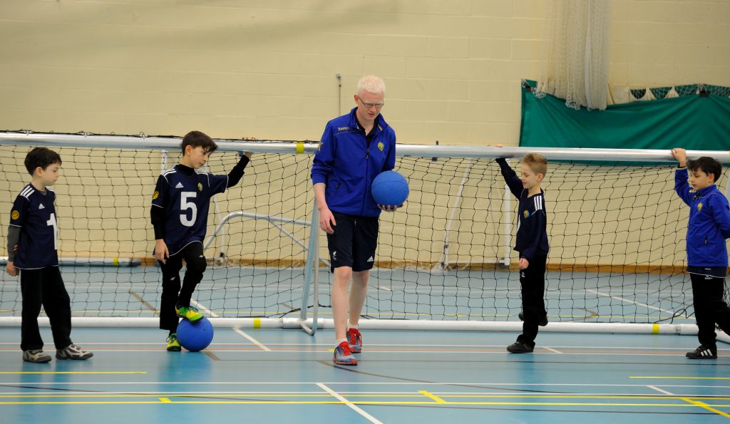Adam holds up a ball in front of a goal with four young goalball players listening to him on the court