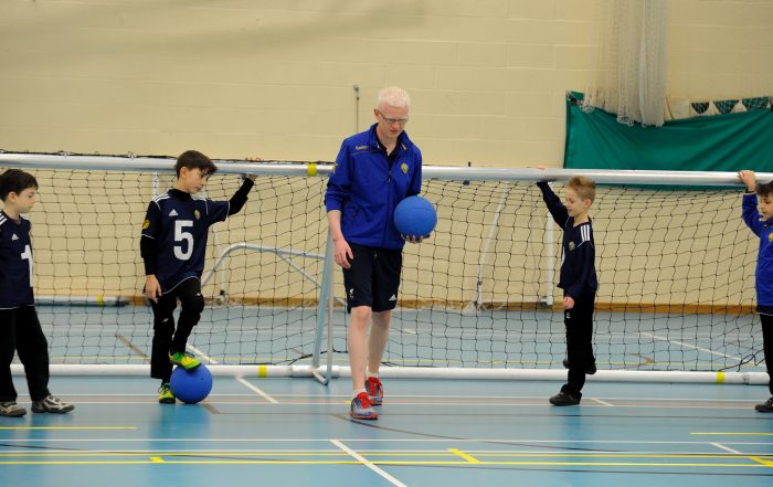 Adam holds up a ball in front of a goal with four young goalball players listening to him on the court