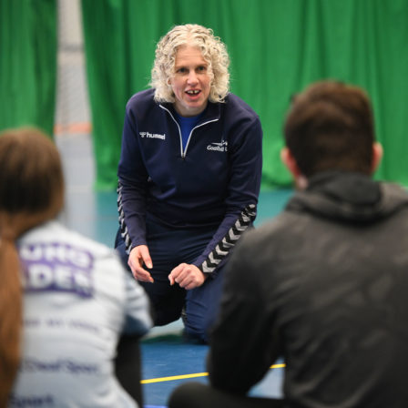 Kathryn Fielding is kneeling on the floor of a sports hall in Goalball UK kit talking to participants of a taster session