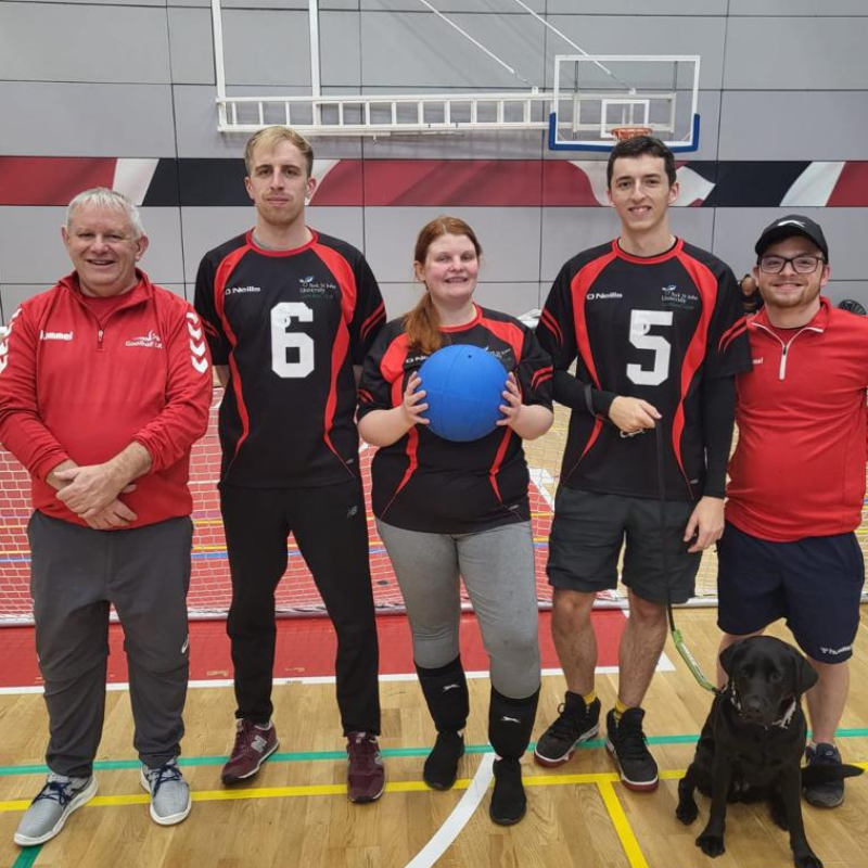 Members of York St John goalball club stand for a photo with the centre player holding a goalball. A guide dog is in the image as well as two coaches