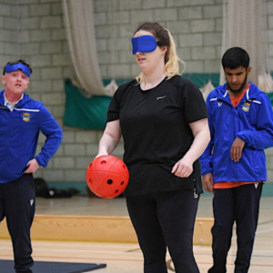 A woman stands in activewear in a sports hall holding a soft goalball and wearing soft eyeshades to try goalball