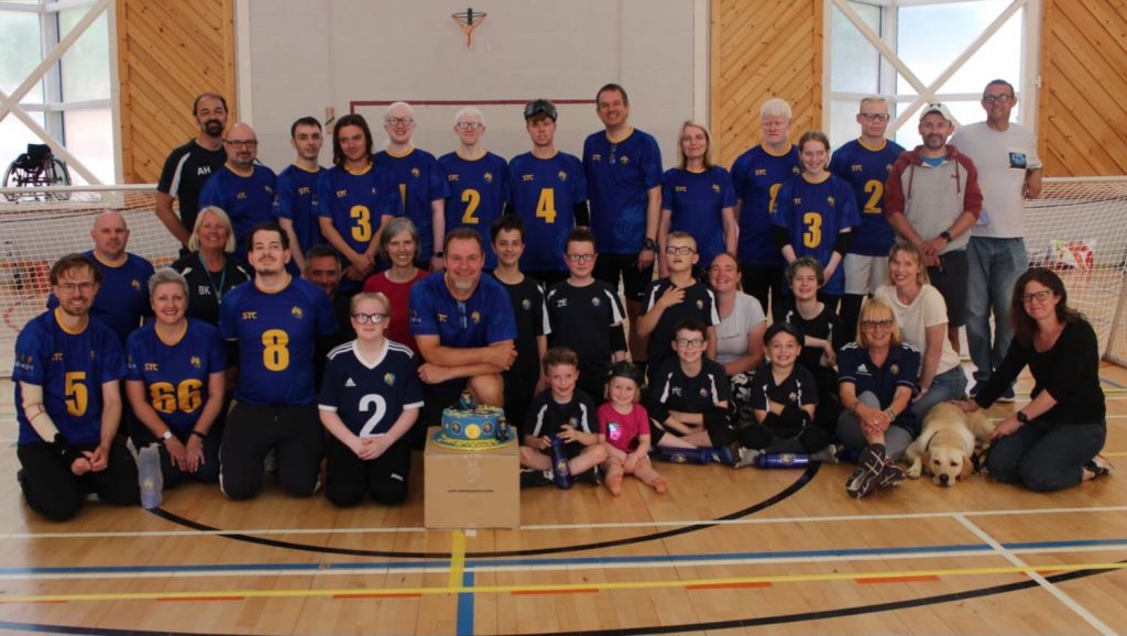 A large group of Winchester Goalball Club members pose around a birthday cake wearing their club kit