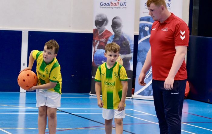 Two young children stand in a sports hall, one of them is holding a soft orange goalball whilst a GB player supervises