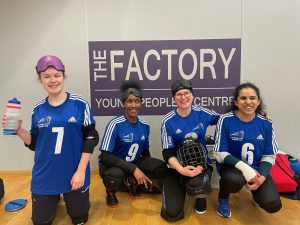 Four female goalball players smile for a photo whilst kneeling on the floor during a break. They are wearing blue tops and their eye shades are on the top of their heads.
