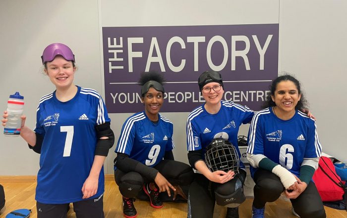 Four female goalball players smile for a photo whilst kneeling on the floor during a break. They are wearing blue tops and their eye shades are on the top of their heads.