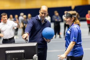Prince William holds a goalball in one hand and points at a monitor with another whilst Lois Turner, GB athlete stands next to him
