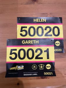 Two race numbers are placed on a wooden surface. They are black, yellow and pink. The top number is 50020 for Helen, and the bottom number is 50021 for Gareth