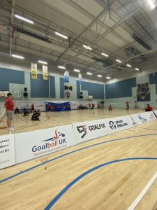 A large sports hall with a goalball court set up with a game in play. In the foreground are a long line of sponsor advertising boards