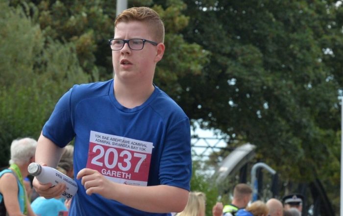 Gareth, a young man with short red hair and black glasses runs in a race wearing a blue t-shirt. He carries a white water bottle and there is a race number 2037 on his t-shirt.
