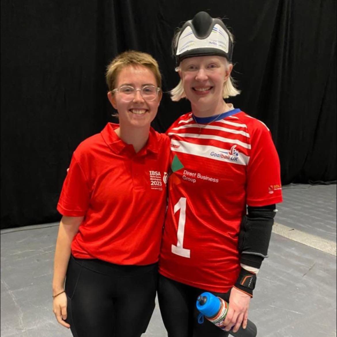 Maggie with short hair and a hearing aid smiles next to an international goalball player